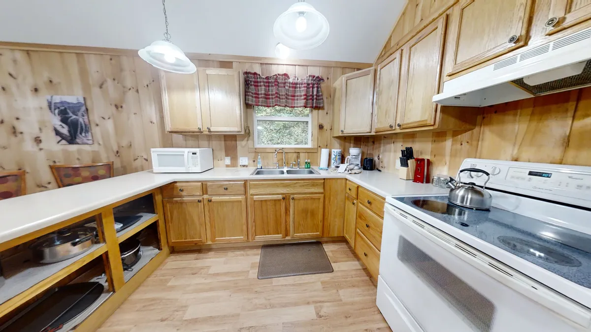 A close-up perspective of the kitchen featuring wooden cabinetry, a kitchen sink, an electric stove, microwave, and a window adorned with a curtain. The inviting setup is ideal for meal preparation, coffee conversations, and family gatherings around the stove.