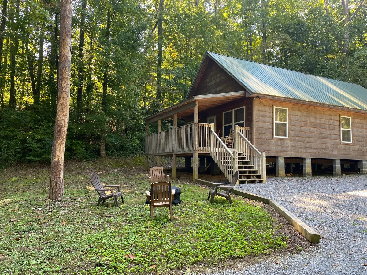 This exterior image showcases the wooden cabin nestled among tall trees, with a welcoming porch featuring outdoor seating and a visible fire pit area. The surrounding natural landscape and serene wooded setting evoke a sense of privacy and tranquility.