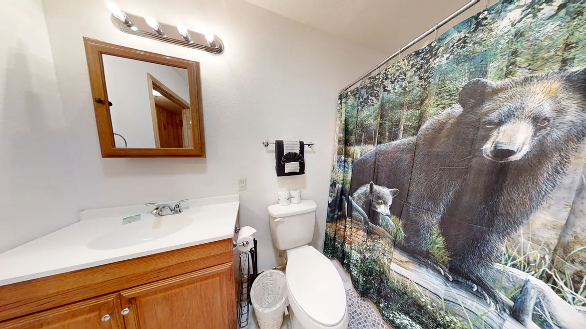 The bathroom features a bear-themed shower curtain, a wooden vanity with a sink and mirror, and warm lighting. The inviting space reflects nature-inspired decor and offers a tranquil setting for daily routines.