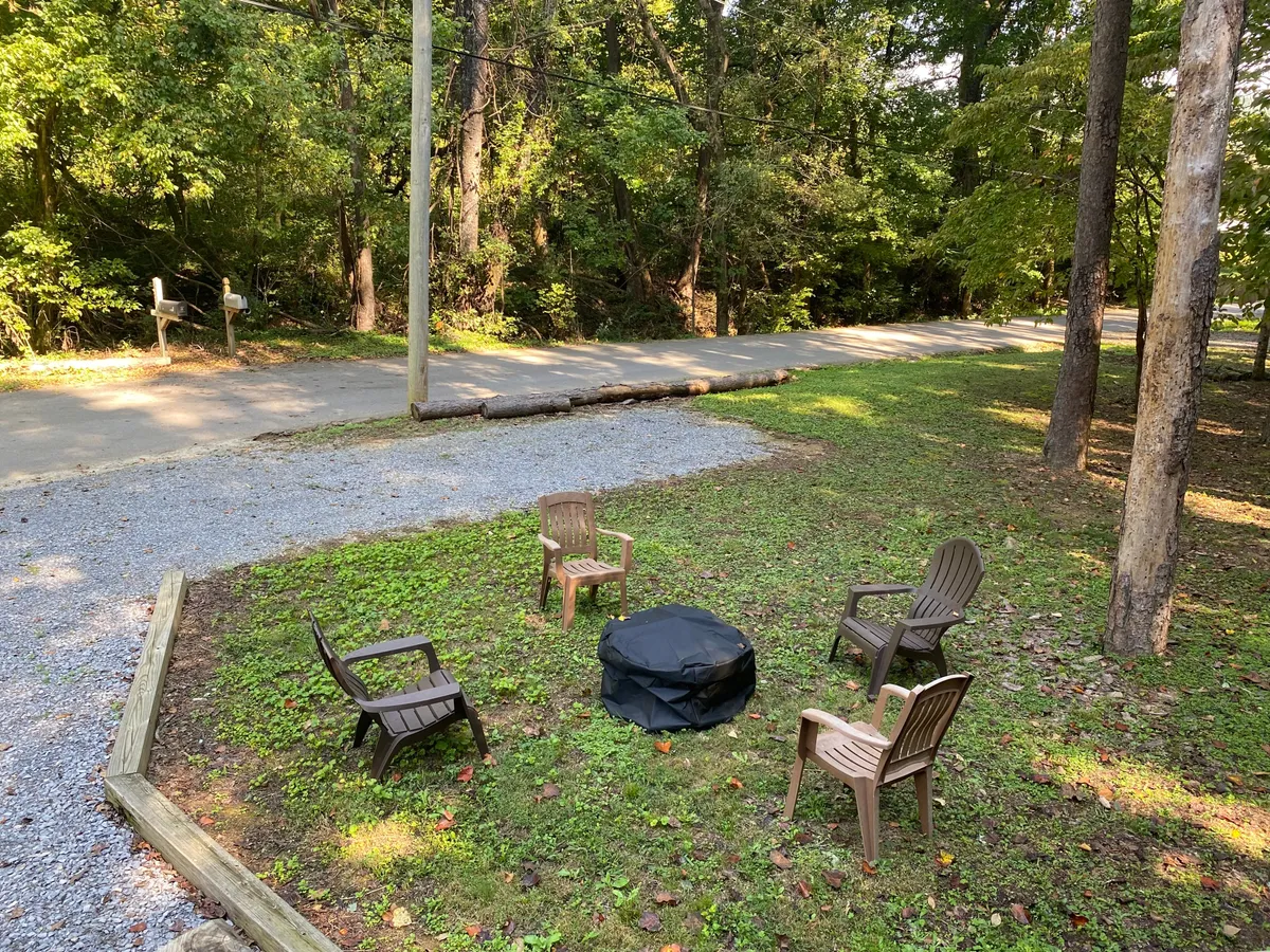 A cozy fire pit area nestled in lush greenery, surrounded by comfortable chairs. The setting is intimate and inviting, perfect for evening gatherings, toasting marshmallows, or quiet conversation under the stars. The abundant trees offer a sense of seclusion and connection to nature.