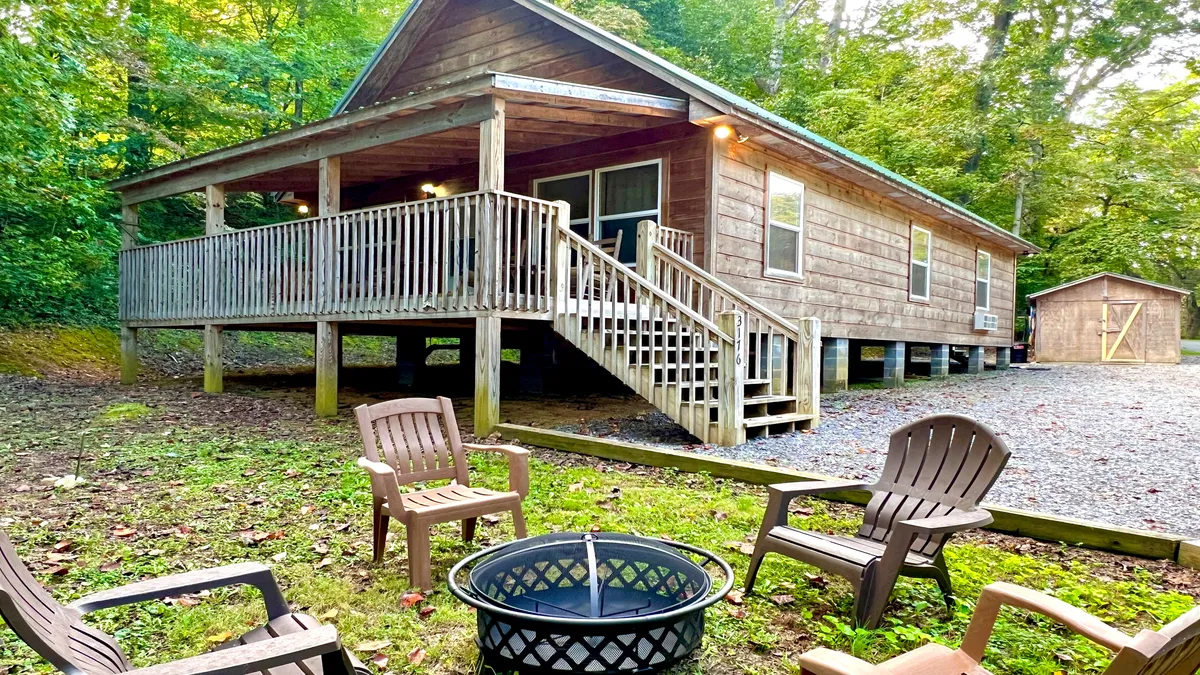 The image showcases a charming, rustic wooden cabin nestled amidst lush greenery in Wears Valley, Tennessee. The foreground features a cozy fire pit surrounded by inviting patio furniture, evoking a sense of warmth and togetherness. The natural setting is tranquil and secluded, ideal for outdoor relaxation and intimate gatherings under the stars.