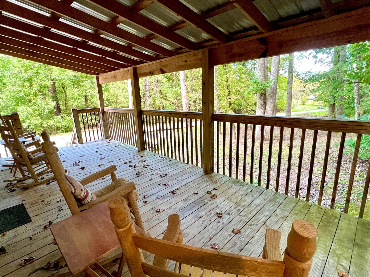 A covered wooden deck with classic rocking chairs overlooks a lush woodland setting. The wooden railing frames the scene, and the area is surrounded by tall trees, offering a tranquil atmosphere. The arrangement invites relaxation, with ample space to enjoy the forest views and the sounds of nature.