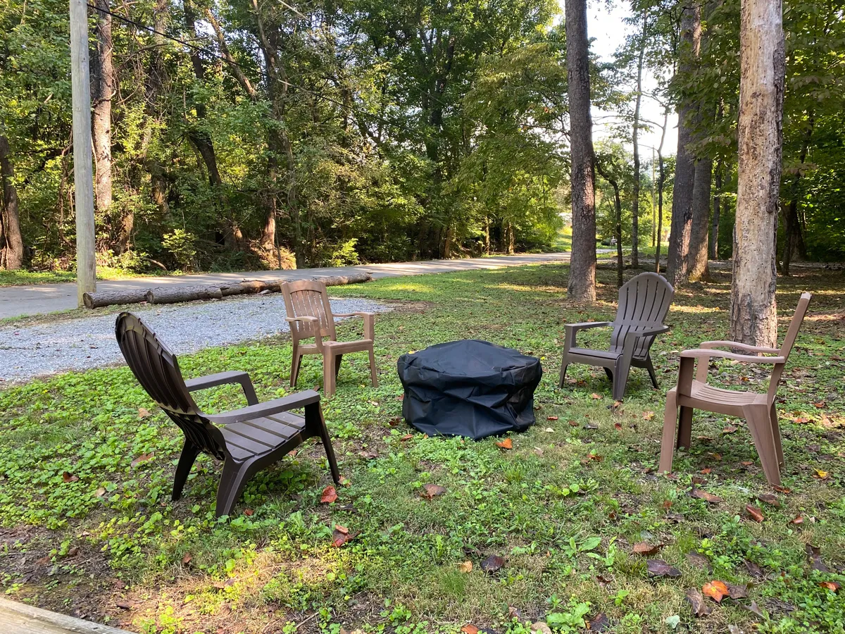 A fire pit covered with a black tarp sits at the center of a flat, grassy yard, surrounded by four brown chairs and bordered by leafy green trees. The open space is ideal for both socializing and letting pets play safely, with nature all around.