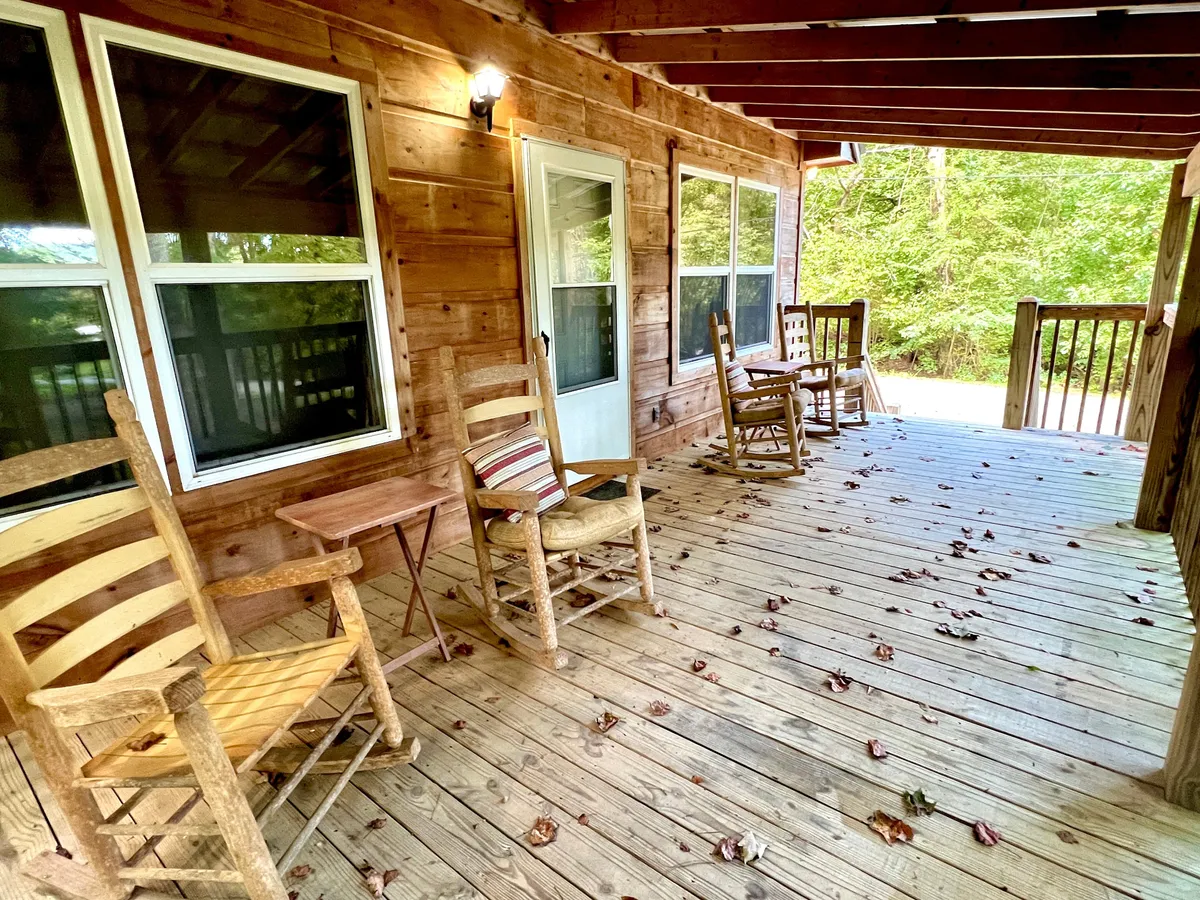 The image highlights two wooden rocking chairs and a table on a spacious deck, with large windows reflecting the forest and soft natural light. The surrounding greenery provides a sense of intimacy and seclusion, inviting couples to relax and connect.