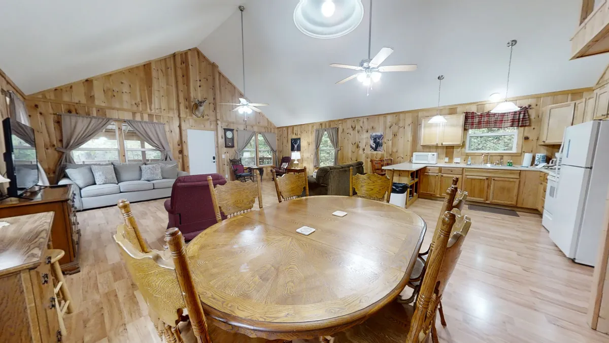 A spacious dining area featuring a large wooden table with seating for six, adjacent to a comfortable seating area and visible kitchen appliances. The space is set up for family meals, lively gatherings, or quiet mornings with coffee and a view of nature.