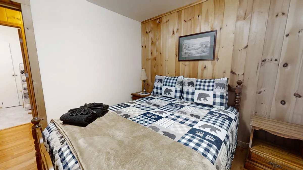 The image focuses on a neatly made bed with patterned bedding, wooden wall paneling, a nightstand with a lamp, and framed artwork, all contributing to a warm and inviting bedroom setting. The soft morning light adds to the sense of peace and comfort.
