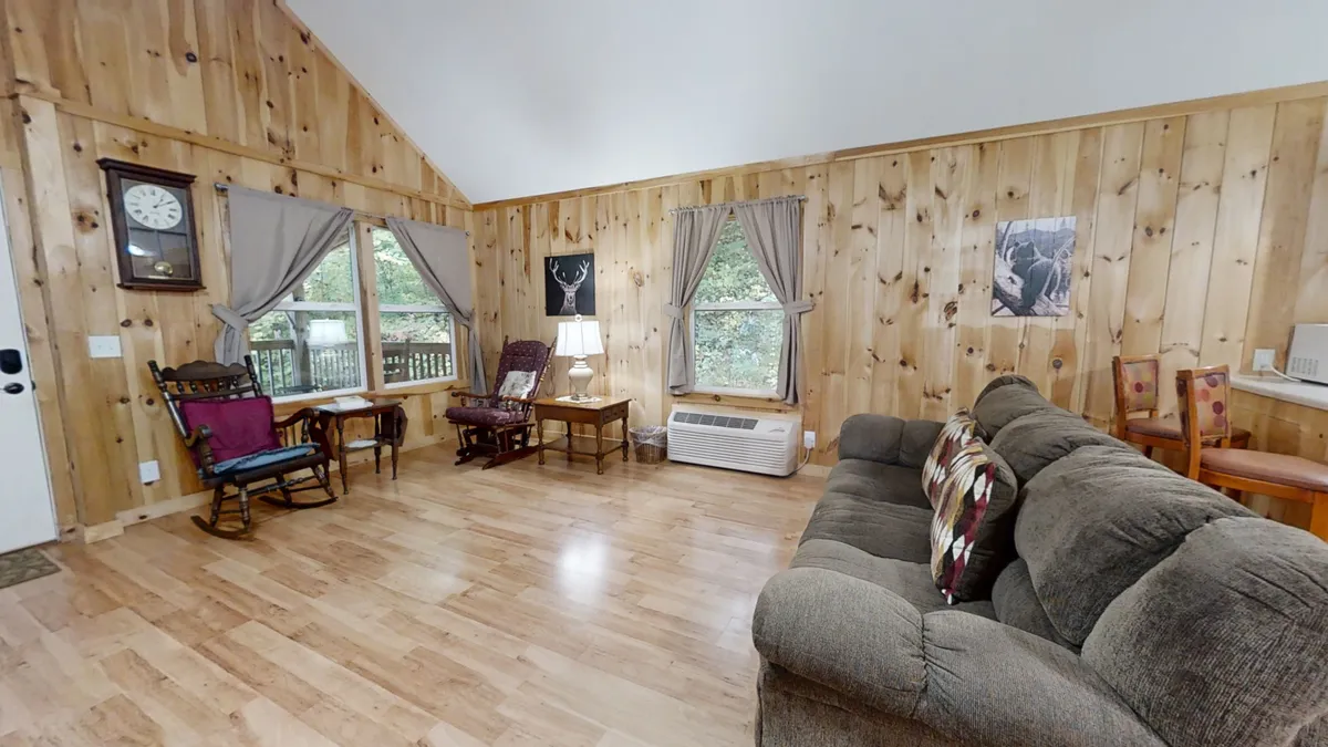 This scene presents another angle of the living area, focusing on a rocking chair, sofa, window with curtains, table lamp, and wooden wall art. The warm ambiance invites relaxation, reading, and sharing stories, with natural light filtering through the window enhancing the peaceful feel.