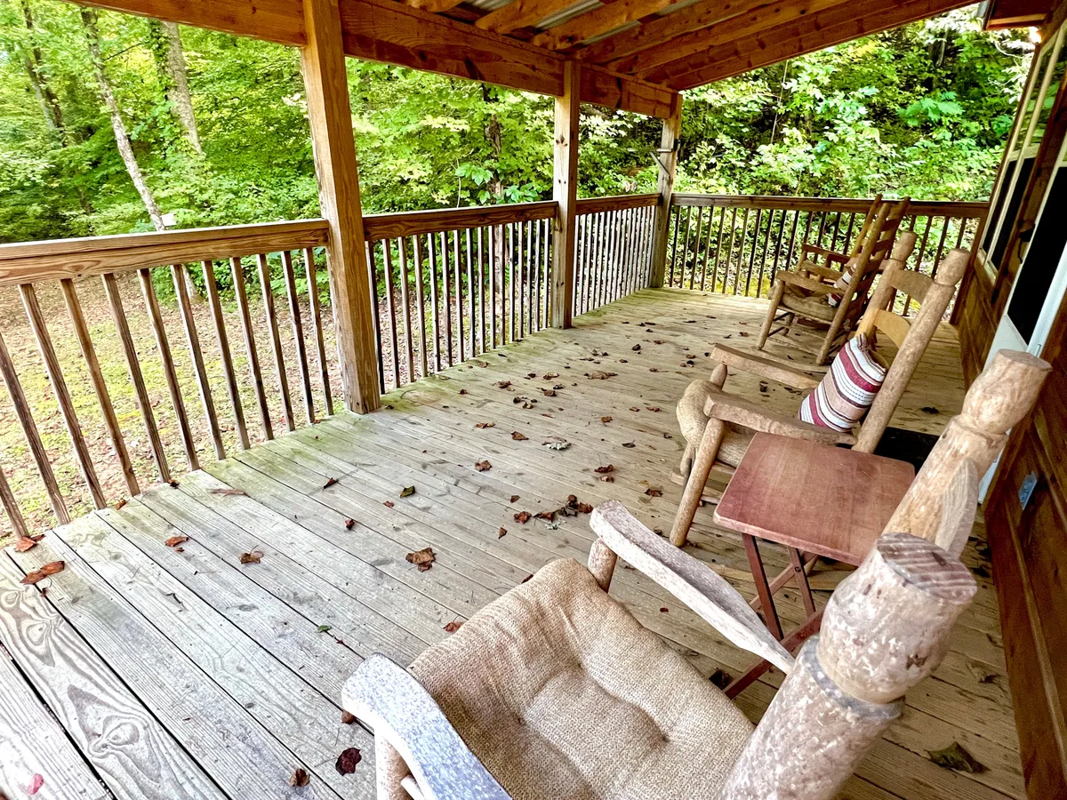 A relaxed deck scene with rocking chairs, rustic wooden railing, and autumn leaves on the floor, offering views into the surrounding woods. The peaceful setting suggests leisurely activities like sipping coffee, reading, or stargazing together.